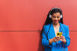 © Dani D.G - Young african woman wearing blue clothes on red background, using her mobile phone, listening to music with wireless headphones. Copy space.