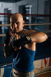 © dikushin - Vertical shot of bald aggressive boxer male wearing bandages punching air to camera in sport club with dark interior, on background of ring. Professional fighter fighting shadow looking at camera.