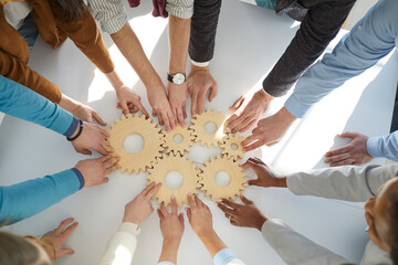 Top view close up of multiracial diverse businesspeople connect cogwheels work together for shared result. Employees join wheel gears involved in teambuilding at workplace. Teamwork concept.