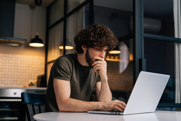 Side view of thoughtful young man programming on laptop sitting at table in kitchen room with modern interior. Bearded pensive male web developer typing script on computer in remote workplace.