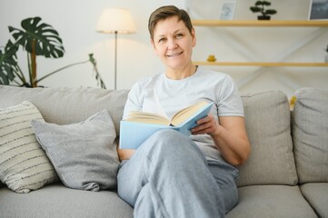 Canvas Print - Middle-aged housewife sitting on sofa with book, free time retirement, hobby.