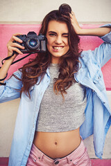 Poster - Enjoy moments and capture memories. Portrait of a beautiful young woman holding a dslr camera and posing against a wall outside.