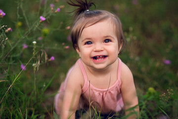 a baby in a pink romper with a ponytail smiles at the viewer from some long grasses with pink flower