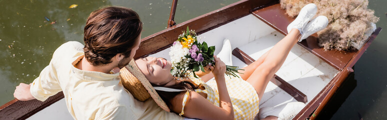 high angle view of young woman in straw hat holding bouquet of flowers and lying in boat with man, banner.