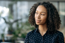 Close-up Of Attractive Smart Young Adult African American Woman With Curly Hair, Manager, Trader, IT Specialist, Standing In A Modern Office, Looking To The Side, Thinking, Dreaming, Smiling