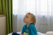 © Marina - A 4-year-old blond-haired boy watches TV while sitting on a bed by the window.
