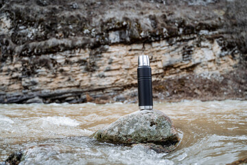 Wall Mural - The concept of a metal vacuum thermos against the background of nature, a thermo bottle stands on a stone in the river, Hiking spring rocks. Tourist utensils.