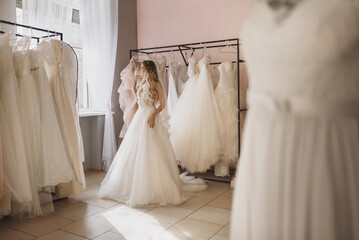 Young blonde woman trying on a dress in wedding salon. A bride with wedding dresses and mannequins in boutique. Full length portrait