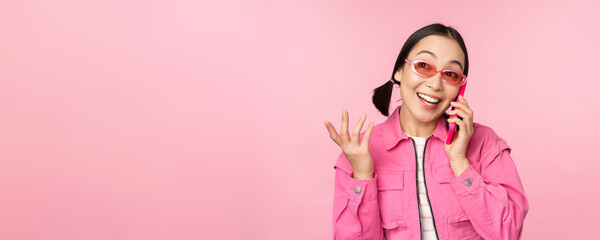 Beautiful korean female model in sunglasses, talking on mobile phone with happy face, using cellular service to call friend on smartphone, standing over pink background