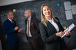 © KAMPUS - Portrait of businesswoman with documents. Happy woman standing in office holding financial reports smiling and looking at camera on male colleagues background. Female leadership in business concept