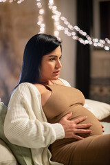 Side view of attractive pregnant woman sitting on bed. Barefoot woman in dress and cardigan touching big belly. Pregnancy, expectation concept