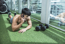 A Fit And Handsome Man Talks To A Friend On The Phone While Lying On The Grass Matting At The Gym. Relaxing In Between Or After Training.