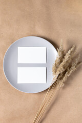 Blank white paper card on plate for mock up with dried spikelets decoration on craft beige background.