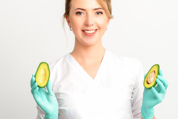 Portrait of smiling young female nutritionist doctor with organic avocado fruits posing at camera on white background, copy space. Benefits of proper nutrition