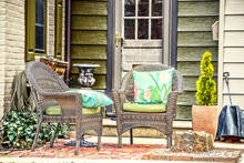 Small Brick Patio On Brick And Rustic Wood Siding House With Two Wicker Chairs With Bright Pillows And Plants And Water Hose Hanging On Wall - Selective Focus