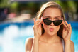Pretty woman making duckface facial expression and holding sunglasses near swimming pool at resort. Girl striking a pose in shades with lips pressed together as in a pout in tropical spa on vacation.
