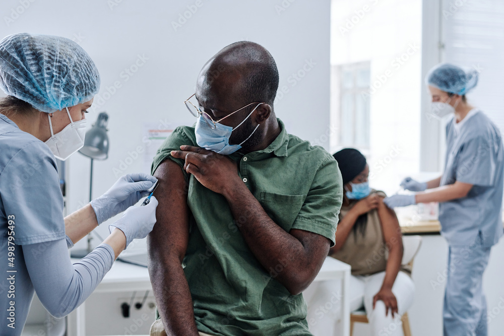 His One Injection African guy in mask preparing his shoulder while doctor giving an ...
