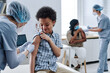 © AnnaStills - African brave boy getting vaccination in shoulder from nurse during procedure at hospital with other people in background