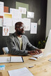 © AnnaStills - African male doctor in suit sitting at his workplace in front of computer monitor and working online at office