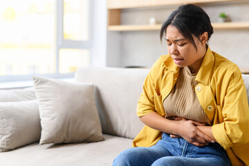 Young African-American woman sitting on the couch and suffers from month menstruation period pain, stomach cramps, multiracial female is holding tummy with a grimace of misery on her face