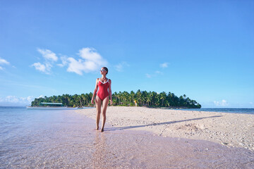 Poster - Vacation on the seashore. Young woman in red swimsuit walking on the beautiful tropical white sand beach.