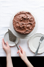 Whole Chocolate Cake Covered In Chocolate Buttercream, On White Cake Stand, With Hands Holding A Vintage Cake Slice.
