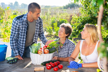 Smiling elderly woman with two adult children breezily chatting at wooden table in garden on summer day happy with rich harvest of vegetables