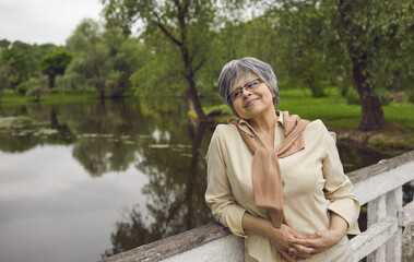 Smiling mature grey-haired woman relax on lake enjoy leisure weekend outdoor. Happy elderly grandmother stand on wooden bridge near river dreaming thinking of good maturity or retirement.