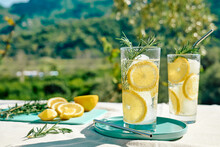 Summer Refreshing Lemonade Drink Or Alcoholic Cocktail With Ice, Rosemary And Lemon Slices On The Table In The Garden. Fresh Healthy Cold Lemon Beverage. Water With Lemon.
