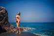 © DSMT - beautiful young woman in a brown swimsuit walks by the beach at the sea with rocks on a summer sunny day. Vacation concept
