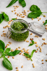 Pesto sauce in a spoon on a glass jar of pesto sauce with ingredients on a white wooden table. Traditional Italian pesto for making fettuccine, pasta, bruschetta. Homemade basil pesto with pine nuts.