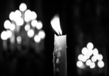 Candles Light. Easter Time Celebtation. Single Candle With Dripping Wax And Blurring Lights Of Many Candles In Two Candlesticks At Background. Black White Photo
