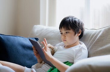 Wall Mural - Young boy using tablet playing game on internet, Kid sitting on sofa watching or talking with friend online,Child relaxing in living room in the morning, Children with New Technology concept