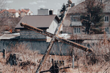 Old Forgotten Burial With Metal Cross At Abandoned Cemetery Against Houses. Old Churchyard. Mystery And Spiritualism Concept. Cemetery Repair. Selective Focus