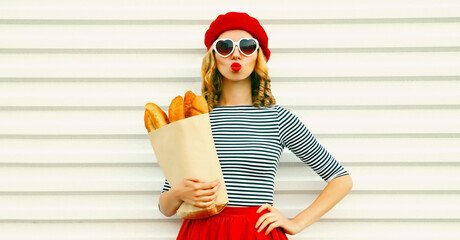 Portrait of beautiful woman blowing her red lips sending sweet air kiss holding grocery shopping paper bag with long white bread baguette wearing french red beret on white background