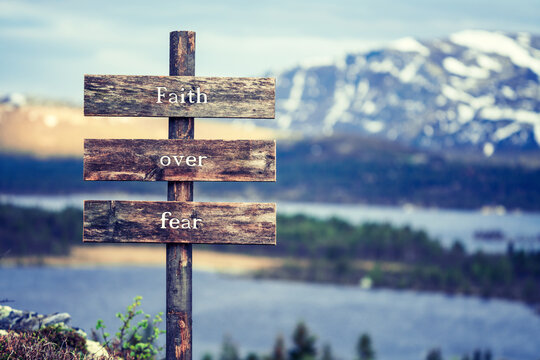 Wall Mural - faith over fear text quote written on wooden signpost outdoors in nature with lake and mountain scenery in the background. Moody feeling.