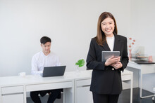 Asian Young Successful Businesswoman Wear Formal Suit Standing And Holding Tablet Smiling With Confident At Office With Her Colleagues Man Working On Laptop On Blur Background. White Background