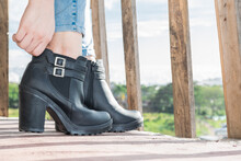 Close-up Detail Of The Feet Of A Latin Girl Putting On Ankle Boots Made Of Black Synthetic Leather, Standing On A Balcony With Wooden Railings, In The Background A Green Rural Colombian Landscape.