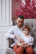 © Tatyana - Handsome happy father sitting with his little daughter on beige sofa indoors. Young man in white shirt with his 1-year-old daughter in white dress, holding her in hands on legs. family tenderness