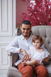 © Tatyana - Handsome happy father sitting with his little daughter on beige sofa indoors. Young man in white shirt with his 1-year-old daughter in white dress, holding her in hands on legs. family tenderness