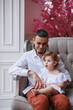 © Tatyana - Handsome happy father sitting with his little daughter on beige sofa indoors. Young man in white shirt with his 1-year-old daughter in white dress, holding her in hands on legs. family tenderness