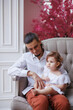 © Tatyana - Handsome happy father sitting with his little daughter on beige sofa indoors. Young man in white shirt with his 1-year-old daughter in white dress, holding her in hands on legs. family tenderness