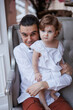 © Tatyana - Handsome happy father sitting with his little daughter on beige sofa indoors. Young man in white shirt with his 1-year-old daughter in white dress, holding her in hands on legs. family tenderness