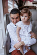 © Tatyana - Handsome happy father sitting with his little daughter on beige sofa indoors. Young man in white shirt with his 1-year-old daughter in white dress, holding her in hands on legs. family tenderness