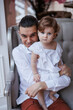 © Tatyana - Handsome happy father sitting with his little daughter on beige sofa indoors. Young man in white shirt with his 1-year-old daughter in white dress, holding her in hands on legs. family tenderness