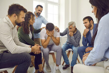 People Comforting A Grieving Crying Man. Concerned Understanding Patients In Group Therapy Supporting A Distraught Desperate Devastated Guy Who's Hiding Face In Hands And Shedding Tears Of Self Pity