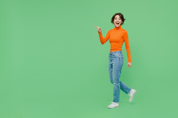 Wall Mural - Full body young smiling happy cool fun woman 20s wearing casual orange turtleneck walk going point index finger aside on workspace area isolated on plain pastel light green color background studio