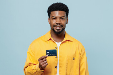 Wall Mural - Young smiling happy fun man of African American ethnicity 20s wear yellow shirt hold in hand credit bank card isolated on plain pastel light blue background studio portrait. People lifestyle concept.