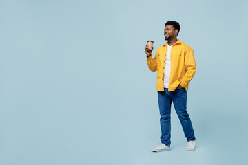 Wall Mural - Full body young man of African American ethnicity 20s in yellow shirt hold takeaway delivery craft paper brown cup coffee to go walking isolated on plain pastel light blue background studio portrait