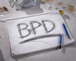 Concept of borderline personality disorder. The abbreviation for BPD is expressively written in pencil with strong pressure on paper. There is crumpled paper and a broken glass on the table.